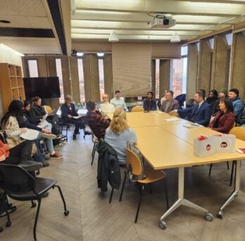 Approximately twenty people sit around a rectangular table, listening to a speaker who is reading from a paper.
                  